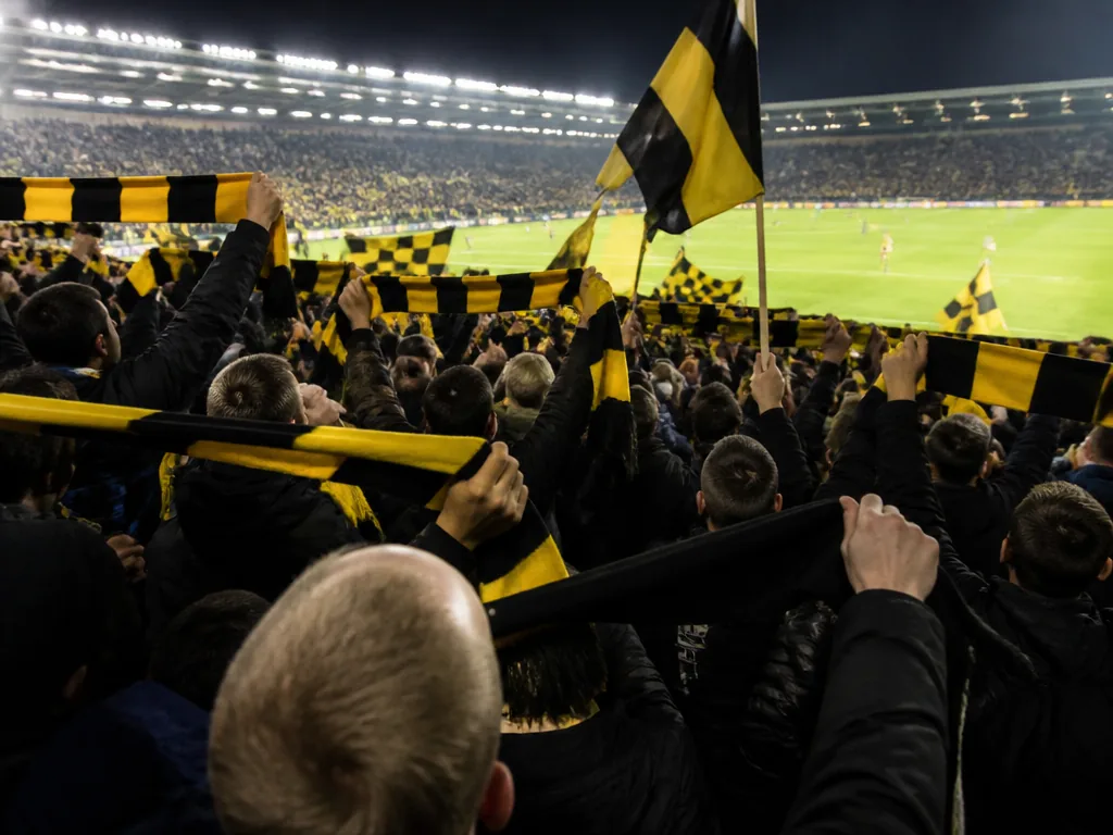 En fylld ståplatssektion i kvällsljus med supportrar som sjunger.